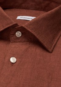 Chemise marron texturée avec col pointu et deux boutons blancs fermés, montrant une étiquette de marque visible à l'intérieur du col.