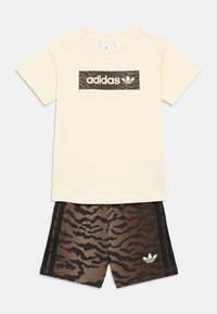 Camiseta Adidas de manga corta en color crema con logo de rayas de tigre en marrón y negro, combinada con pantalones cortos a juego con rayas de tigre y franjas negras.