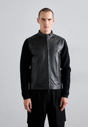 Black leather vest with a zip front and stand collar, contrasted by black knit long sleeves. Features side pockets and a smooth texture.