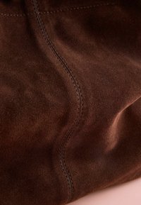 Brown suede bag featuring a soft texture, prominent stitching, and a slightly structured form. Surface exhibits natural variations.