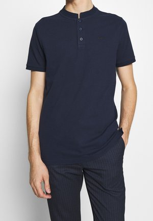 Mężczyzna ubrany w granatowy krótki rękaw henley z trzema guzikami i granatowe spodnie w paski, stojący z jedną ręką w kieszeni.