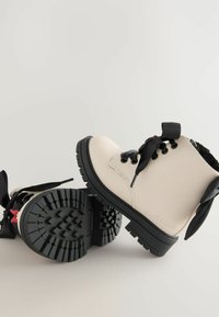 Helle beige Knöchelstiefel mit schwarzen Schnürsenkeln und Gummisohlen. Verfügt über eine strukturierte Sohle für besseren Halt und einen kleinen roten Akzent an der Ferse.