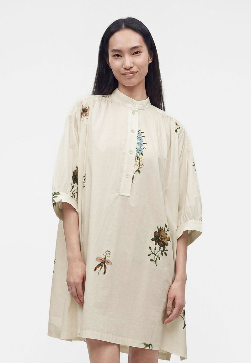 Γυναίκα με μακριά σκούρα μαλλιά φοράει φαρδύ, κρεμ φόρεμα με σχέδια λουλουδιών και εντόμων και στέκεται μπροστά σε απλό φόντο.