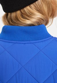 Prošívaná modrá bunda s žebrovaným límcem. Tkanina má hladkou texturu s diamantovým vzorem prošívání a kontrastními detaily švů.