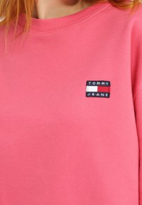 Sweat-shirt rose avec un col côtelé, comportant un patch rectangulaire bleu foncé avec "TOMMY JEANS" en lettres blanches et un accent rouge.