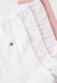 Τέσσερα διπλωμένα καλτσάκια σε λευκούς και ροζ τόνους· μία λευκή κάλτσα έχει ένα μικρό κεντημένο κόκκινο, λευκό και μπλε λογότυπο κοντά στη μανσέτα.