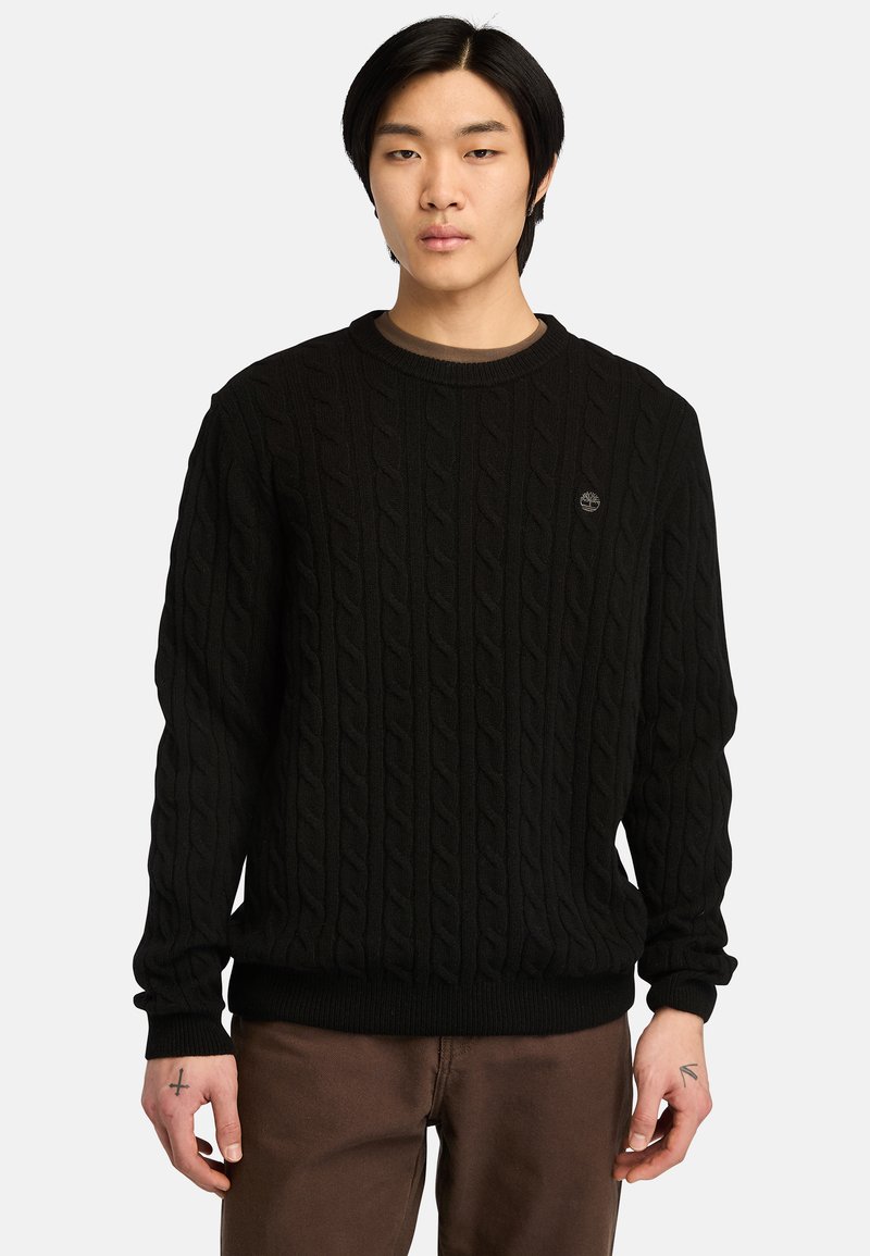 Maglione nero a trecce con colletto rotondo, polsini e vita a costine. Presenta un piccolo logo sul petto. Indossato con pantaloni marroni.