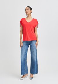 Rode top met korte mouwen, een V-hals en strikdetails aan de mouwen, gecombineerd met wijde blauwe jeans en zilveren puntige schoenen.