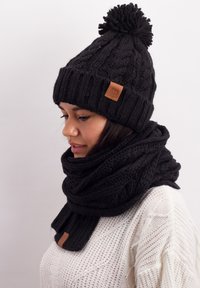 Black cable-knit beanie with a pom-pom and leather label, paired with a matching black scarf featuring a thick, textured pattern.