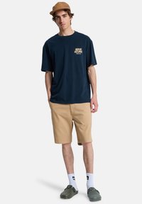 Μπλε ναυτικό t-shirt με λογότυπο και κείμενο, καφέ βερμούδα, λευκές κάλτσες με μαύρο λογότυπο και πράσινα παπούτσια τύπου slip-on. Καθημερινό, άνετη εφαρμογή.