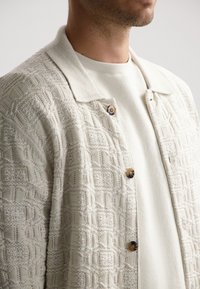 Teksturni kremni cardigan z geometrijskimi vzorci, ki ima obročasto ovratnico in rjave gumbe. Pod njim je vidna svetla pulover.
