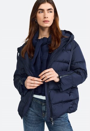 Giacca a vento blu navy con finitura strutturata, dotata di cappuccio, chiusure con zip e bottoni, e tasche laterali con zip. Indossata sopra un maglione blu navy.
