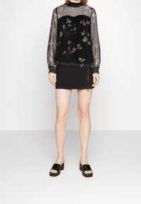 Blouse longue à manches longues en tissu transparent noir à fleurs, avec poignets smockés, associée à une mini-jupe noire dotée d'une fermeture éclair latérale. Mannequin portant des sandales à plateforme noires.