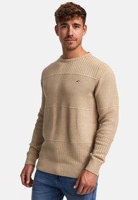 Pull en tricot beige avec des panneaux texturés, doté d'un col rond et de poignets côtelés. Petit logo sur la poitrine.