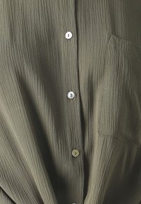 Camicia verde oliva con tessuto testurizzato, caratterizzata da pieghe verticali, chiusura a bottoni e una singola tasca sul petto. Bottoni argentati in dettaglio.