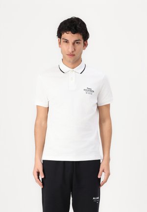 Polo blanc à manches courtes, avec un col noir contrastant et un petit logo brodé sur la poitrine. Associé à un pantalon noir.