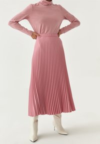 Růžová skládací midi sukně s vysokým pasem, spárovaná s dlouhým rolákem. Modelka nosí bílé boty na podpatku. Hladká textura.