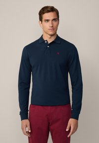 Marineblå langermet polo skjorte med krage, to knapper og en liten rød logo på brystet. Kombinert med røde bukser.