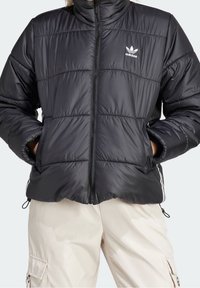 Svart puffjacka med hög krage, quiltad design, blixtlås och vitt Adidas-logga på vänster bröst. Strukturerad tyg.