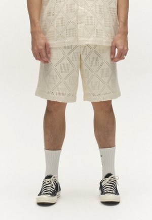 Egy személy alsó testrésze, aki krémszínű, geometriai mintás kötött rövidnadrágot és inget visel, fehér zoknival és fekete alacsony szárú tornacipővel.