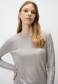 Frau mit langen blonden Haaren, die ein silbernes Langarmoberteil trägt, steht mit einer Hand an der Hüfte vor einem einfarbigen hellen Hintergrund.