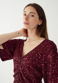 Bordo blizgučiai marškiniai su giliais V formos iškirptėmis, trumpomis rankovėmis ir tekstūruotu audiniu su raukinių dizainu, suteikiančiu papildomą detalumą.