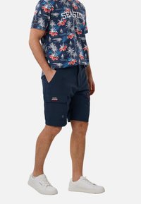 Homme portant un short cargo bleu marine et un t-shirt bleu à fleurs avec l'inscription "SEASIDE", ainsi que des baskets blanches, debout les mains dans les poches.