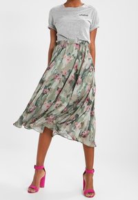 Jupe midi florale en tissu opaque avec un motif camouflage vert et des fleurs roses. Associée à un t-shirt gris et des sandales à talons roses.