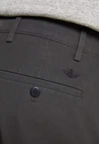 Donkergrijze broek met een achterzakje met een zwarte knoop, subtiele stikseldetails en een geborduurd embleem dat op vleugels lijkt.