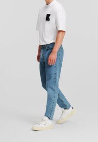 Camiseta blanca de manga corta con parche de logo negro, jeans de mezclilla azules y zapatillas blancas con suela beige. Atuendo simple y casual.