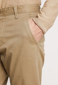 Mano nella tasca anteriore di pantaloni beige indossati con un top beige a maniche lunghe abbinato, con texture del tessuto e dettagli di cucitura visibili.