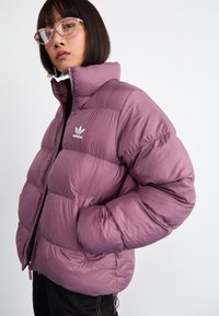 Nafúkaná fialová bunda s vysokým golierom, zipsovým uzatváraním a bielym logom Adidas. Má bočné vrecká a hladký, lesklý povrch.
