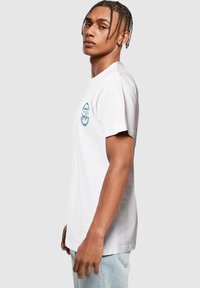 White cotton t-shirt with short sleeves, featuring a blue graphic design on the left chest. Simple round neckline, relaxed fit.