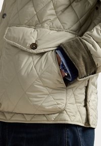 Beige quilted jacket with corduroy accents; features a button closure and visible pocket holding a wristwatch. Dark jeans visible at the bottom.