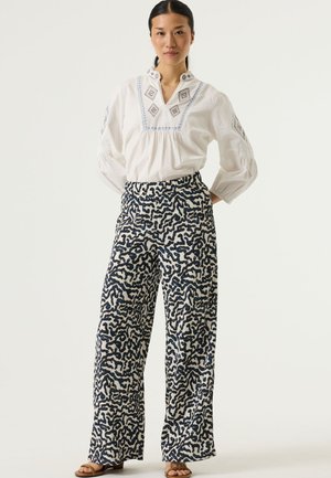 Blusa blanca bordada con un cuello decorativo, combinada con pantalones de pierna ancha de patrón en tonos azul marino y crema. Sandalias que completan el conjunto.