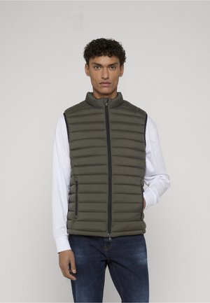 Mężczyzna w oliwkowozielonej pikowanej kamizelce bez rękawów na białej koszuli z długim rękawem i niebieskich dżinsach, stojący na jednolitym jasnym tle.