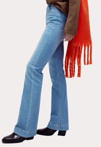 Vaqueros acampanados de un azul claro con cintura alta, que presentan un detalle de costura contrastante y una textura ligeramente desgastada. Combinados con botas de tacón marrones y una bufanda de flecos naranja.