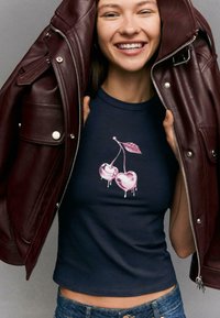 Top corto blu navy con grafica di ciliegie gocciolanti rosa, abbinato a una giacca di pelle bordeaux con dettagli in metallo argentato.