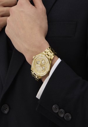 Goldene Uhr mit achteckigem Ziffernblatt, mit Diamantakzenten und einem Metallarmband. Getragen an einem Handgelenk, das teilweise über einem dunklen Anzug mit Knopfdetails sichtbar ist.
