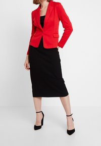 Blazer rouge sur mesure avec revers, robe midi noire côtelée et chaussures à talons noirs avec brides à la cheville. Design minimaliste, silhouette ajustée.