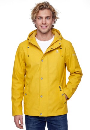 Junger Mann mit welligem blondem Haar, trägt eine gelbe Kapuzen-Regenjacke und blaue Jeans, lächelt und steht mit einer Hand in der Hosentasche.