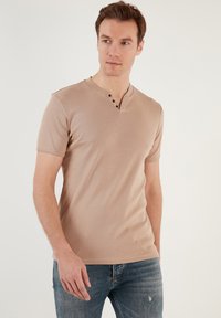Krátký béžový tričko s výstřihem do V a třemi knoflíky v přední části. Měkká látka a přiléhavý střih. Nosené s modrými džíny.