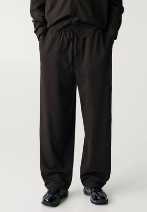 Persoon draagt losse zwarte broek met elastische tailleband en trekkoord, zwarte schoenen en een deels zichtbaar zwart bovenstuk met de handen in de zakken.