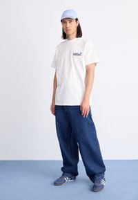 White cotton t-shirt with logo on left chest, paired with loose blue denim jeans and grey athletic shoes featuring white stripes and textured detail.