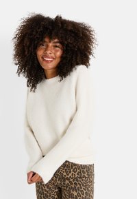 Suéter de punto color crema con un acabado texturizado, cuello redondo y mangas largas, combinado con jeans de estampado de leopardo.