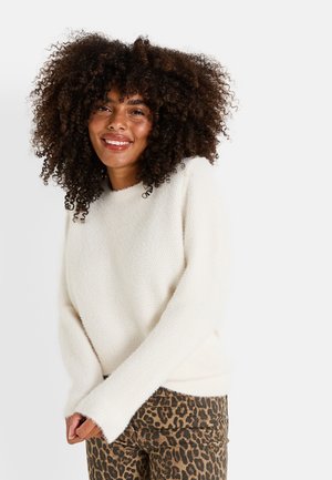 Suéter de punto color crema con un acabado texturizado, cuello redondo y mangas largas, combinado con jeans de estampado de leopardo.