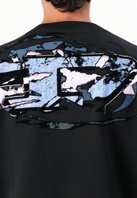 Personne portant un t-shirt noir avec un grand design graphique abstrait en bleu et rose, usé, sur le haut du dos.