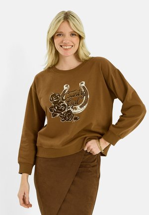 Femme blonde souriante portant un sweat marron avec un fer à cheval, des roses et le texte "Country Girl", assorti à une jupe marron.