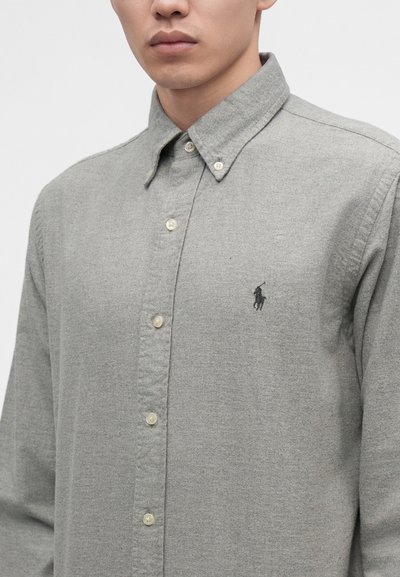 Άνδρας που φοράει γκρι ανοιχτό πουκάμισο με κουμπιά, με κουμπωμένο γιακά και μικρό κεντητό σκούρο λογότυπο στο αριστερό στήθος.