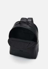 Schwarzer Leder-Rucksack mit doppelten Schultergurten, einer blickdichten Innentasche mit Reißverschluss und einem Stofffutter, das den Markennamen trägt.